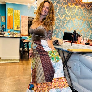 May include: A woman wearing a brown velvet top and a long, multi-colored patchwork skirt with a white ruffle trim. The skirt is made of different fabrics and patterns, including floral, paisley, and plaid. She is standing in a room with blue walls and a patterned wallpaper.