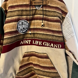 May include: A brown, green, and white striped hoodie with a patch that says "Widespreed Panic" and the text "Aint Life Grand" on the front. The hoodie has a drawstring hood and a kangaroo pocket.