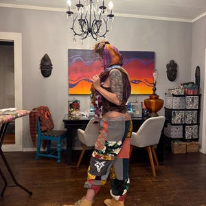 Pu&ograve; includere: Una persona indossa pantaloni patchwork con motivi floreali e geometrici, abbinati a stivali da lavoro color cuoio. La persona indossa anche una sciarpa colorata e un top scuro. La stanza ha un lampadario e opere d'arte sulla parete.