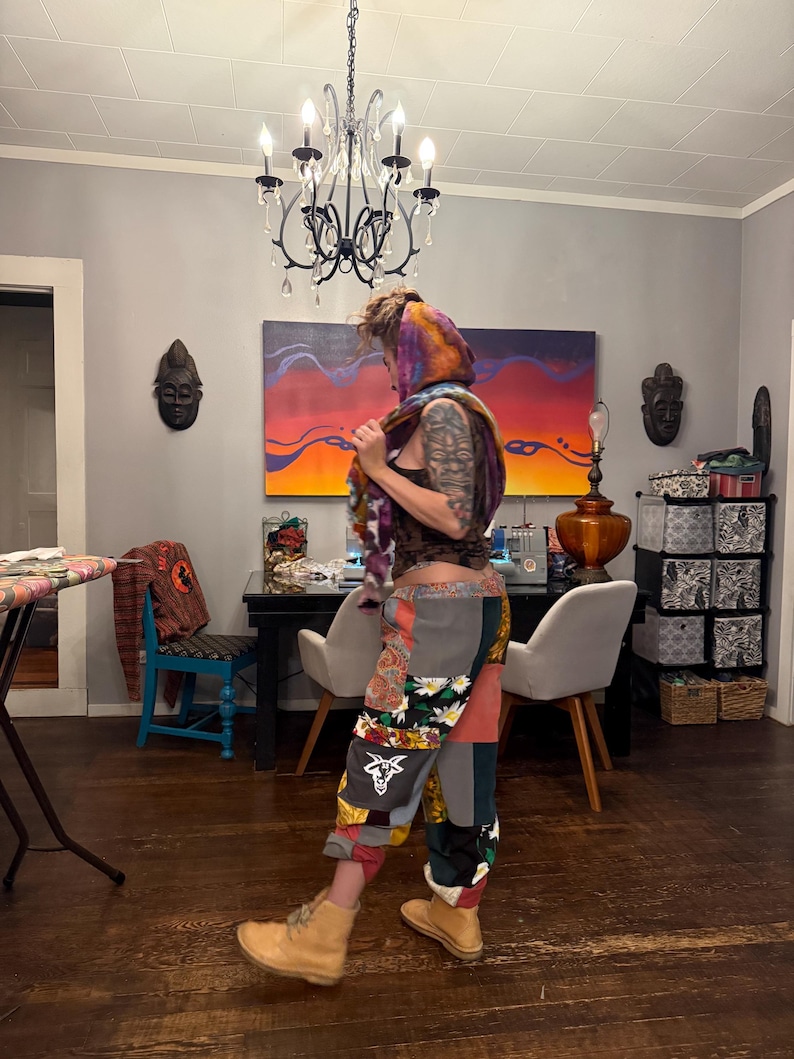 Pu&ograve; includere: Una persona indossa pantaloni patchwork con motivi floreali e geometrici, abbinati a stivali da lavoro color cuoio. La persona indossa anche una sciarpa colorata e un top scuro. La stanza ha un lampadario e opere d'arte sulla parete.