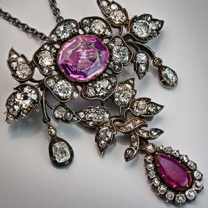 Könnte beinhalten: Eine verzierte Anhängerkette mit floralem Design. Die Kette hat einen großen, ovalen, rosa Edelstein in der Mitte, umgeben von kleineren, klaren Edelsteinen. Ein tropfenförmiger Rubin hängt darunter, ebenfalls von klaren Steinen umgeben.