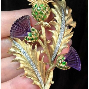 May include: A gold thistle brooch with carved purple amethyst flower heads and green gemstone detailing. The brooch has a detailed leaf design with a line of small, clear gemstones, set against a black background.