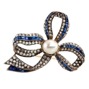 May include: A vintage bow brooch with a central pearl and intricate detailing. The brooch features a silver-toned metal design with clear and blue gemstones, creating a sparkling effect. The bow shape is a classic design, suitable for formal wear.