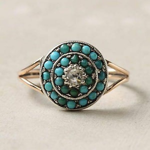 Puede incluir: Un anillo dorado con un diseño circular. El anillo presenta una piedra central transparente rodeada por dos círculos concéntricos de cuentas turquesas. La banda se divide en dos en la parte superior, creando un aspecto elegante.