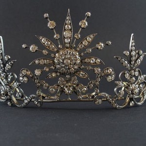May include: A detailed silver tiara featuring a central sunburst motif, embellished with numerous sparkling gemstones. The tiara showcases elaborate floral and scrollwork patterns, presenting a majestic and refined look. The gemstones are set in silver metal.