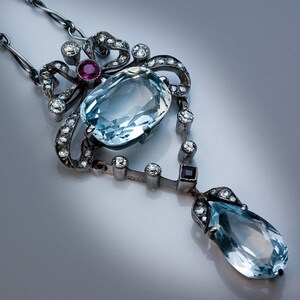 Op de afbeelding: Een antieke zilveren ketting met een grote, ovale, lichtblauwe edelsteen, geaccentueerd met kleinere diamanten en een druppelvormige edelsteen. Een kleine, ronde, rode edelsteen is ook aanwezig. De ketting heeft een sierlijk, vintage ontwerp.