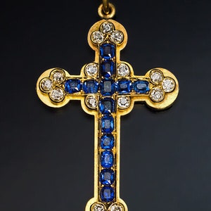 Op de afbeelding: Een gouden kruisvormige hanger met een centrale kolom van vierkante, diepblauwe edelstenen. Het kruis is verder versierd met witte edelstenen en een gouden zetting. De hanger hangt aan een zwart koord.