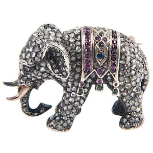 May include: Silver elephant brooch with detailed gemstone embellishments. The elephant has a decorative saddle with purple, blue, and green gemstones. The brooch features a pin closure on the reverse.