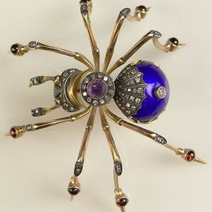 Puede incluir: Broche de araña dorada con abdomen de esmalte azul, adornada con diamantes y una amatista central. Las patas de la araña son doradas con detalles de granate. Joya detallada.