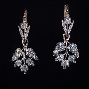 May include: A pair of elegant drop earrings featuring a floral design. Each earring has a gold-tone hook and a silver-tone setting adorned with numerous sparkling, clear gemstones. The earrings are set against a black background.