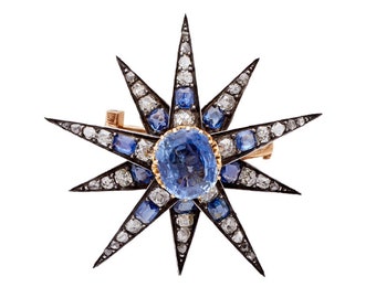 Könnte beinhalten: Sternförmige Brosche mit einem zentralen ovalen Saphir, umgeben von kleineren blauen und weißen Edelsteinen. Die Goldfassung und die schwarzen Emaille-Details bilden einen auffälligen Kontrast. Die Sternspitzen sind scharf und detailliert.