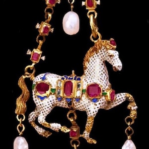 Puede incluir: Colgante dorado con un caballo esmaltado blanco con detalles rojos y azules, adornado con piedras preciosas de color rubí y perlas. El caballo cuelga de una cadena decorativa con gemas adicionales y una gran gota de perla.