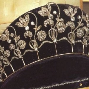 May include: A detailed tiara featuring a floral pattern, adorned with many glittering gemstones. The silver-coloured tiara sits on a dark velvet surface inside a wooden box. A matching bracelet is also visible.