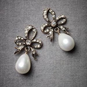 May include: A pair of bow-shaped earrings with a gold-toned metal design. The earrings are adorned with clear crystals and have a pear-shaped, white pearl pendant. The earrings are displayed on a gray, textured background, showcasing their intricate details.