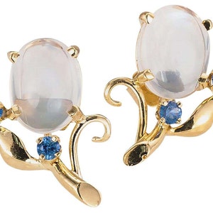 May include: A pair of gold earrings with large, oval, clear gemstones. Each earring has a floral design with blue gemstones and a curved stem. The earrings are set against a white background.