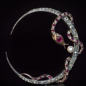 May include: A silver-toned brooch in the shape of a stylized snake encircling a circle. The snake's head features a red gemstone eye and a pearl. The snake's body is adorned with small, sparkling stones and red gemstones. The brooch is set against a black background.