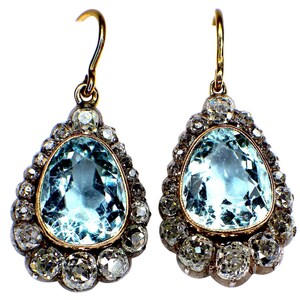 May include: A pair of drop earrings with large, pear-shaped, pale blue gemstones. Each earring is framed by a halo of smaller, clear gemstones, set in a gold-coloured metal. The earrings have a classic, vintage design, suitable for special occasions.