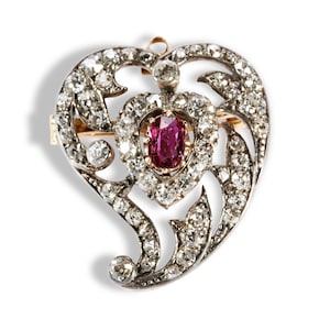 Könnte beinhalten: Eine herzförmige Brosche mit einem zentralen Rubin, umgeben von Diamanten. Die Brosche hat ein durchbrochenes Design mit zusätzlichen Diamanten, die im Filigran gesetzt sind. Die Fassung scheint aus Gold und Silber zu sein.
