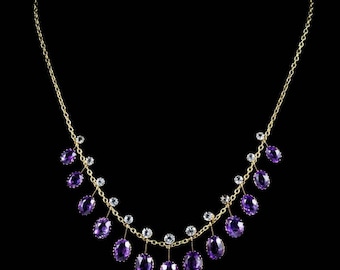 May include: A gold-toned necklace with a delicate chain design. The necklace features oval-shaped, deep purple gemstones and small, clear gemstones. The gemstones are evenly spaced along the chain, creating a symmetrical and elegant look. The background is black.