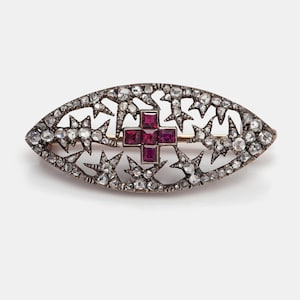 May include: An antique brooch with an oval shape, featuring a central cross design made of square-cut red gemstones. The brooch is surrounded by a filigree pattern of stars and clear gemstones, set in a silver-toned metal.