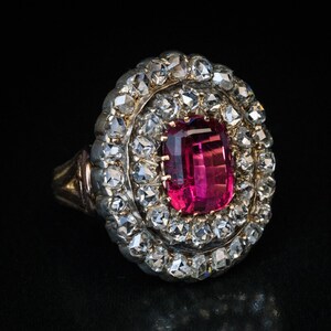 May include: A vintage ring with a large, rectangular, deep pink gemstone at its centre, encircled by two halos of small, clear gemstones. The ring's setting is gold-toned with detailed design, set against a black backdrop.