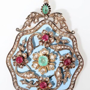Op de afbeelding: Een sierlijke hanger met een lichtblauwe emaille achtergrond, met bloemmotieven met rode en groene edelstenen. De goudkleurige lijst is versierd met heldere stenen en een decoratieve top met een groene edelsteen.