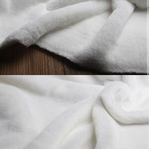 May include: White faux fur fabric with a soft, plush texture. The fabric is folded and draped, showcasing its luxurious feel.