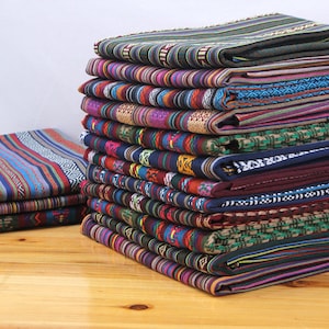 May include: A stack of colorful fabric with various patterns and stripes. The fabric is woven with different colors, including blue, red, green, yellow, and purple. The fabric is folded neatly and stacked on a wooden surface.
