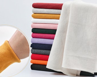 Könnte beinhalten: Ein Stapel farbiger Rippstoffe in verschiedenen Farbtönen, darunter Rot, Grau, Gelb, Weiß, Rosa, Grün und Lila. Ein separates Stück weißen Stoffes ist zur Seite drapiert. Nahaufnahme eines gelben Rippstoffärmels.