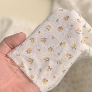 May include: Close-up of a white fabric with a repeating pattern of small yellow and gray floral designs. The fabric is held up, showing its texture and the details of the floral print. The fabric appears to be lightweight.