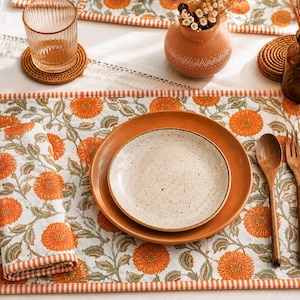 Può includere: Una tovaglietta rettangolare con motivo floreale arancione su sfondo bianco e bordo a righe. Include un tovagliolo piegato abbinato, un piatto grande color terracotta, un piatto più piccolo maculato e posate in legno.