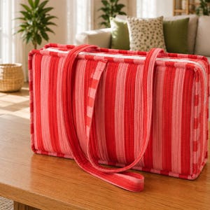 May include: A rectangular quilted tote bag featuring vertical stripes in various shades of red and pink. The bag has a white zipper closure along the top edge and two long fabric shoulder straps with a red and pink checkered pattern.