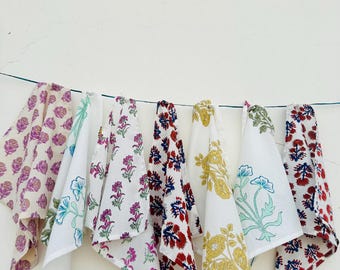 Pode incluir: Uma coleção de sete lenços de algodão branco, cada um adornado com um padrão floral único em várias cores. Os desenhos incluem roxo, azul, vermelho e dourado, criando uma exibição vibrante. Os lenços estão pendurados em um fio fino contra uma parede branca.