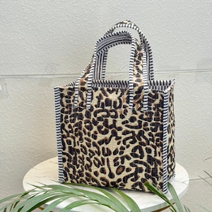 May include: A quilted tote bag featuring a brown and black leopard print pattern on the main body. The bag has two top handles and side panels decorated with a black and white vertical striped fabric trim, providing a structured rectangular shape.