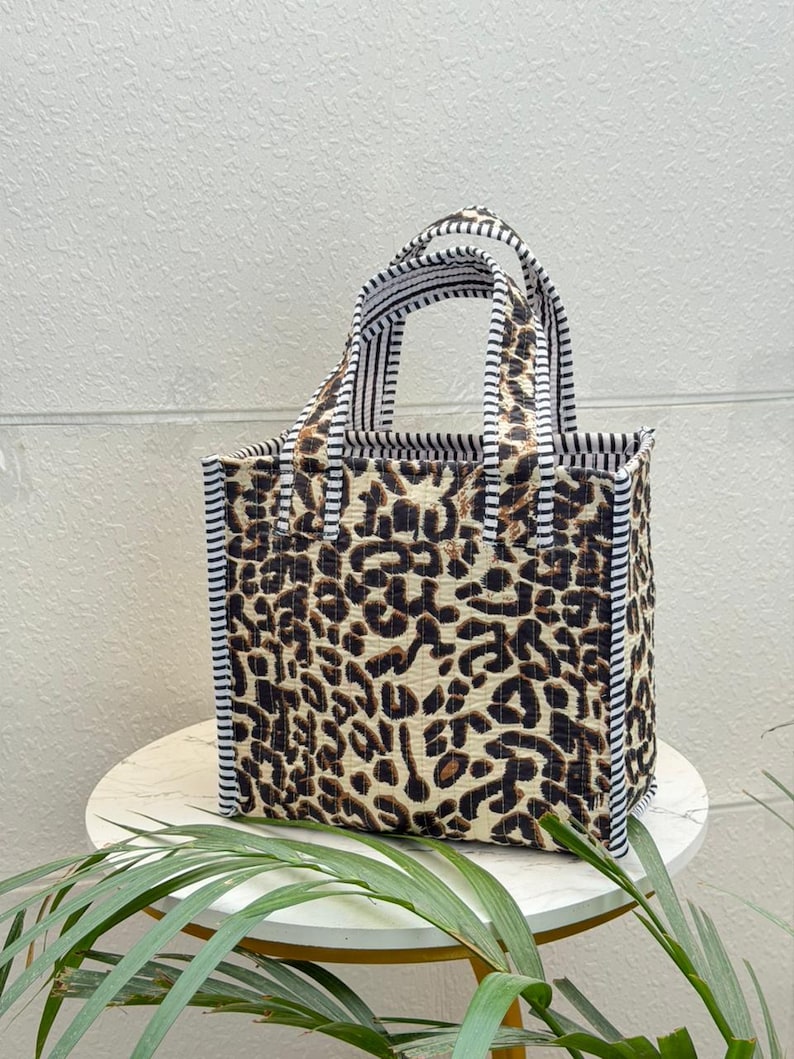 May include: A quilted tote bag featuring a brown and black leopard print pattern on the main body. The bag has two top handles and side panels decorated with a black and white vertical striped fabric trim, providing a structured rectangular shape.