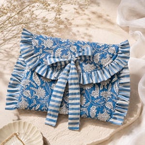 May include: A quilted fabric pouch featuring a blue and white floral pattern with a ruffled edge and a blue and white striped tie closure. The rectangular pouch has a flap that secures with two long fabric ribbons tied in a bow at the centre.