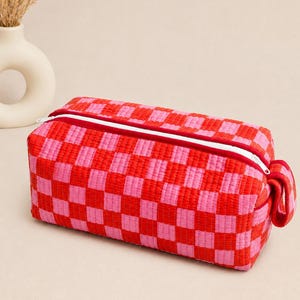 May include: A rectangular quilted pouch featuring a vibrant red and pink checkered pattern. The bag has a white zipper closure along the top and a small fabric loop on the side. The quilted texture is visible across the entire surface of the pouch.