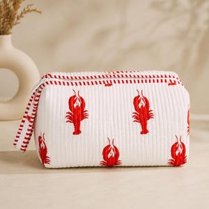 May include: A white quilted rectangular toiletry bag featuring a repeating pattern of bright red lobsters. The bag has a zipper closure with a white and red striped fabric pull tab and matching striped trim along the top edge and side seams.
