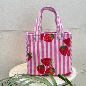 May include: A quilted tote bag featuring a pattern of vertical pink and white stripes with a repeating strawberry print. The bag has two matching striped fabric handles attached to the top, a rectangular base, and a structured upright shape.