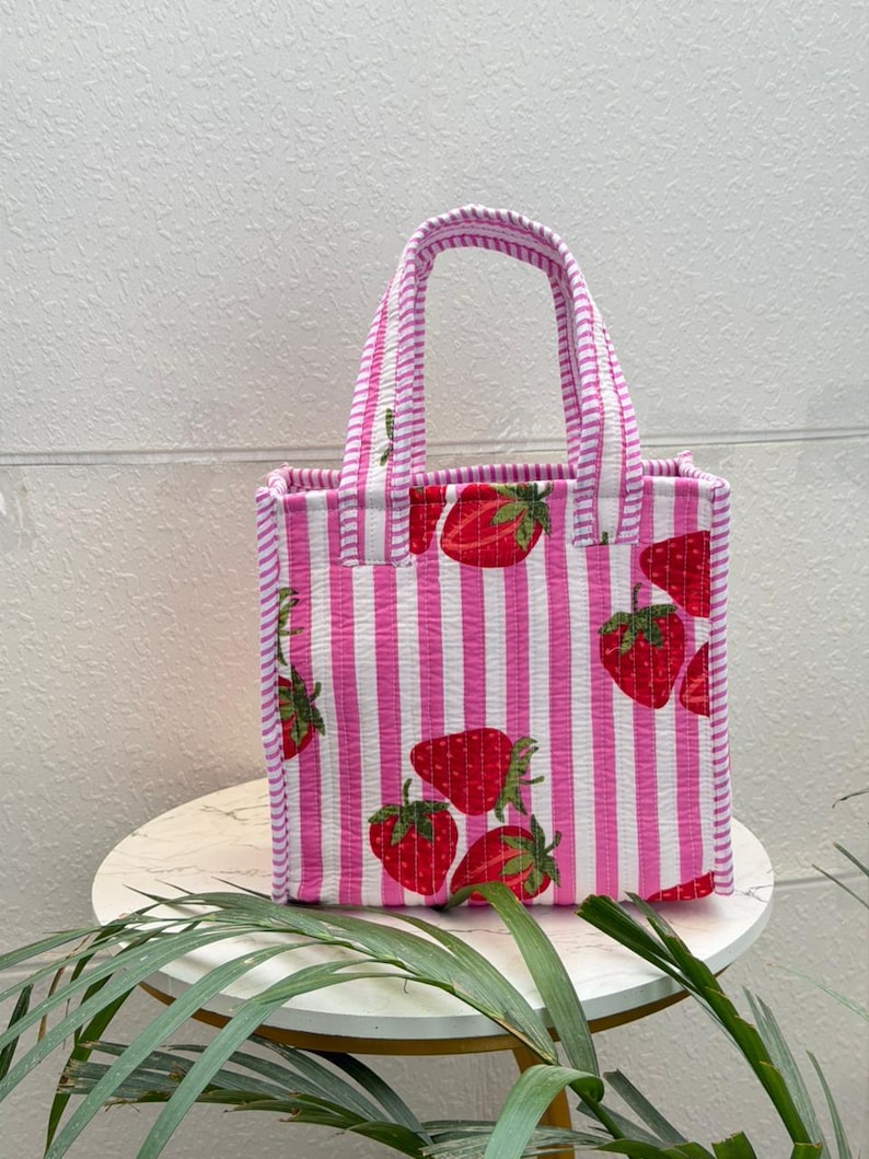 May include: A quilted tote bag featuring a pattern of vertical pink and white stripes with a repeating strawberry print. The bag has two matching striped fabric handles attached to the top, a rectangular base, and a structured upright shape.