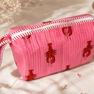 May include: A quilted pink zippered pouch featuring a repeating pattern of red lobsters. The pouch has a rectangular shape with a matching pink and white striped fabric trim along the zipper line and a coordinating striped fabric wrist strap attached.