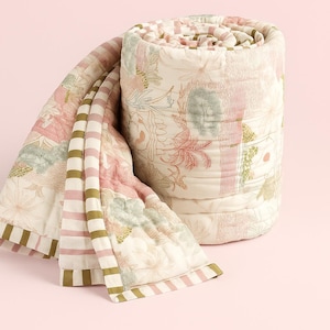 May include: Rolled-up floral quilt with a cream background and pink, green, and brown floral patterns. The quilt has a striped border in pink and olive green. The quilt is rolled up and partially unrolled, showing the detailed stitching.