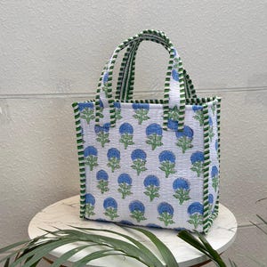 May include: A quilted tote bag featuring a white base with a repeating pattern of blue flowers and green stems. The bag has two top handles and vertical side edges decorated with a green and white striped fabric trim, creating a structured look.