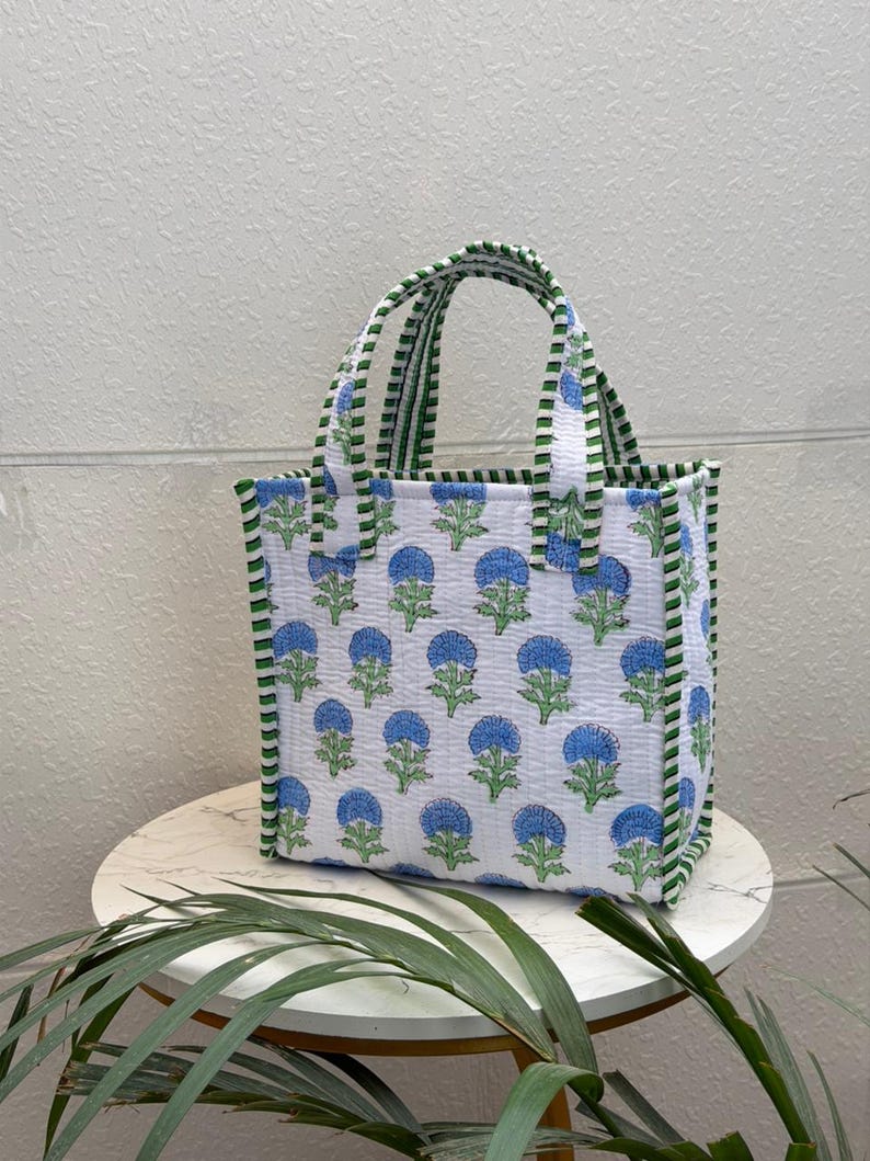 May include: A quilted tote bag featuring a white base with a repeating pattern of blue flowers and green stems. The bag has two top handles and vertical side edges decorated with a green and white striped fabric trim, creating a structured look.
