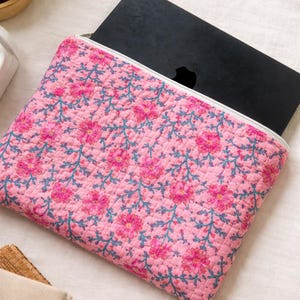 May include: Pink floral laptop sleeve with a white zipper. The quilted fabric features a repeating pattern of pink flowers and teal vines. A black laptop is partially visible inside the sleeve.