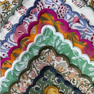 Könnte beinhalten: Eine Sammlung geschichteter Stoffservietten mit gewellten Rändern und bestickten Säumen in verschiedenen Farben wie Senfgelb, Dunkelgrün, Blau, Magenta und Weiß. Jede Serviette zeigt ein florales Muster mit botanischen Motiven.