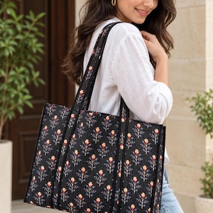 May include: A black quilted tote bag featuring a repeating floral pattern with red, yellow, and blue flowers on thin green stems. The bag has two long black shoulder straps and a rectangular structure with visible vertical stitching throughout the fabric.