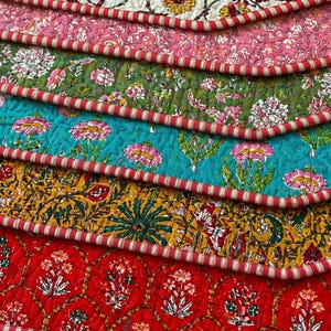 May include: A collection of patterned, quilted fabric pieces in a variety of colours. The layers include white, pink, green, teal, yellow, and red, each with a different floral design. The edges are trimmed with a red and white striped border.