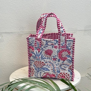 May include: A quilted tote bag featuring a white base with a floral pattern in shades of pink and blue. The bag has two handles and side panels decorated with a pink and white striped fabric trim that matches the interior lining of the bag.
