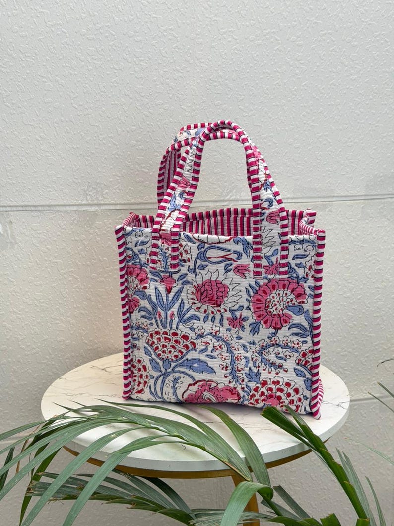 May include: A quilted tote bag featuring a white base with a floral pattern in shades of pink and blue. The bag has two handles and side panels decorated with a pink and white striped fabric trim that matches the interior lining of the bag.